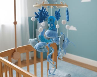 May include: A baby mobile featuring felt sea creatures in shades of blue, including a seahorse, jellyfish, and starfish. The mobile hangs from a wooden ring and is suspended above a crib. White pom-poms and a blue rug complete the nursery scene.