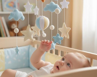 May include: A baby in a crib gazes up at a felt mobile. The mobile features a sun, clouds, stars, and a hot air balloon in shades of blue, yellow, and white. The crib is made of light wood and has blue and yellow bedding.