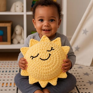 May include: A smiling baby holds a large, yellow, crocheted sun-shaped pillow. The pillow has a smiling face with closed eyes, made with black yarn. The baby is wearing a gray long-sleeved shirt and gray pants.