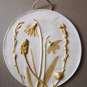 May include: A round, white ceramic wall hanging with a gold-painted design of flowers and leaves. The design is raised and textured, giving it a 3D effect.