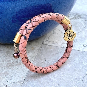 May include: A braided pink cork bracelet featuring two gold-toned metal end caps. One end cap holds a gold-toned hibiscus flower charm, while the other end cap features a small dangling dark purple pearl bead with a clear crystal accent.