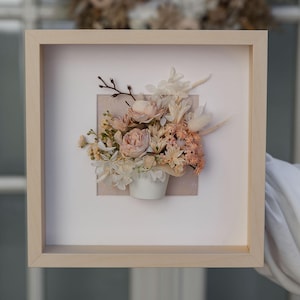 Può includere: Un arrangiamento di fiori pressati incorniciato con una varietà di fiori secchi in tonalità di rosa e bianco, presentato in un piccolo vaso bianco. I fiori sono disposti a grappolo e sono circondati da uno sfondo bianco.