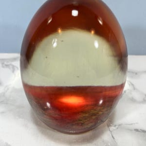 May include: A smooth, egg-shaped decorative object made of clear resin. The top section is a translucent amber colour, while the bottom section features a textured, layered landscape design in deep red, brown, and orange tones with a bright light.