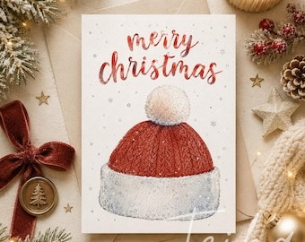 Peut inclure: Carte de Noël avec une illustration à l'aquarelle d'un bonnet de Père Noël rouge avec un pompon et une bordure blancs. Les mots "merry christmas" sont écrits en rouge au-dessus du chapeau. Décorations festives.