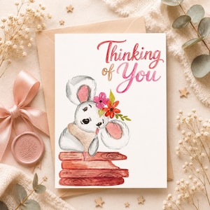 Op de afbeelding: Een wenskaart met een aquarel illustratie van een muis met grote oren, versierd met een bloemenkrans, rustend op een houten constructie. De kaart toont de tekst "Thinking of You" in roze.