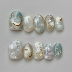 May include: Ten oval-shaped false nails with a cloudy white and light blue base. Some nails feature gold crescent moons and stars, while others have gold stars or glitter.