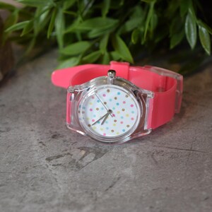 May include: A pink and clear wristwatch with a white face and colorful polka dots. The watch has a clear plastic case and a pink silicone band.