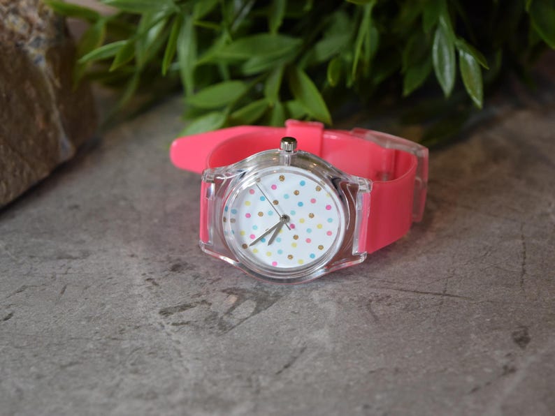May include: A pink and clear wristwatch with a white face and colorful polka dots. The watch has a clear plastic case and a pink silicone band.