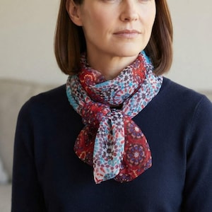 May include: A lightweight scarf featuring a repeating geometric mandala-inspired pattern in shades of deep red, bright blue, and white. The fabric is tied in a simple knot at the neck, displaying a vibrant, symmetrical design across the entire surface.