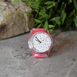 May include: A pink wristwatch with a white face and colorful polka dots. The watch has a clear plastic band.