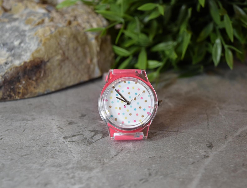 May include: A pink wristwatch with a white face and colorful polka dots. The watch has a clear plastic band.