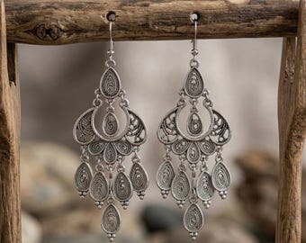 May include: A pair of silver chandelier earrings. Each earring features an ornate design with multiple tiers of filigree and teardrop-shaped dangles. The earrings are suspended from a wooden display stand, showcasing their intricate details and bohemian style.