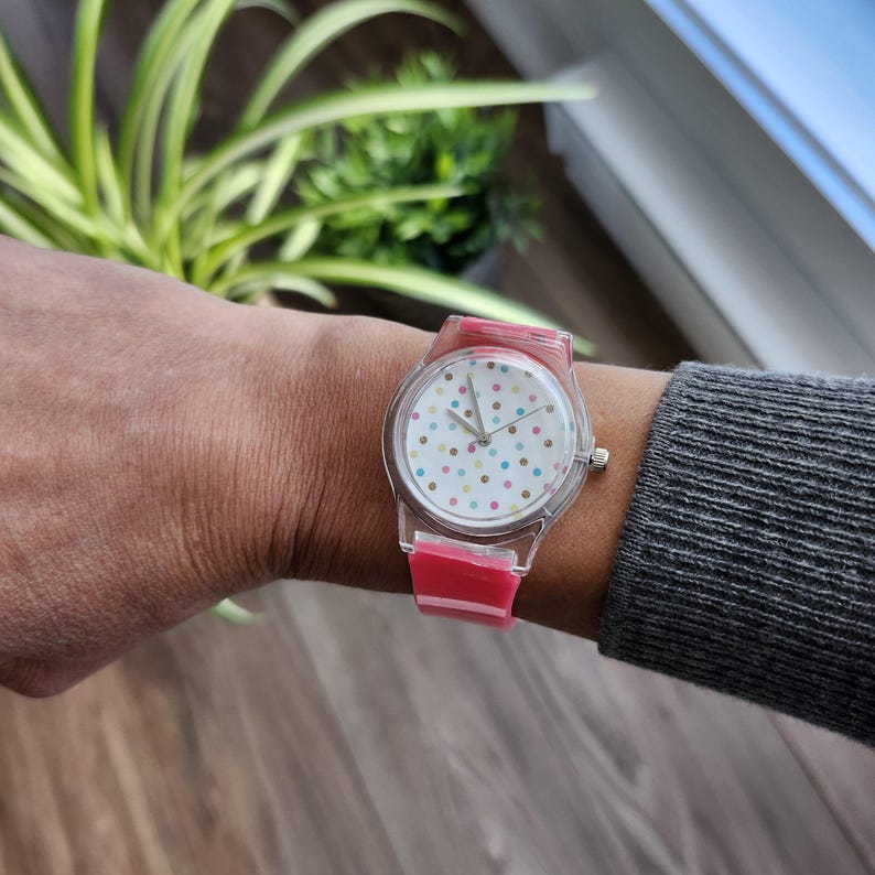 May include: A clear plastic watch with a pink band and a white face with colorful polka dots.