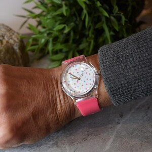 May include: A pink and white wristwatch with a clear plastic band and a white face with colorful polka dots.