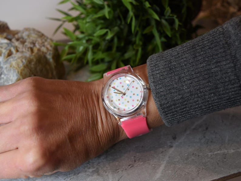 May include: A pink and white wristwatch with a clear plastic band and a white face with colorful polka dots.