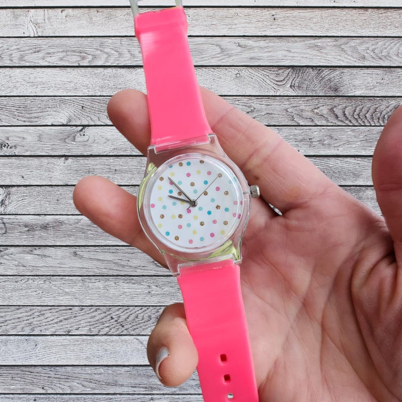 May include: A pink silicone wristwatch with a clear plastic case and a white face with colorful polka dots.
