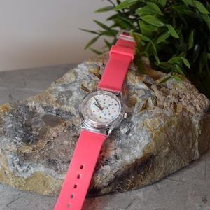 May include: A pink silicone wristwatch with a white face and colorful polka dots. The watch is sitting on a rock.