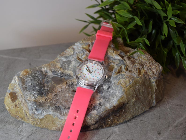 May include: A pink silicone wristwatch with a white face and colorful polka dots. The watch is sitting on a rock.