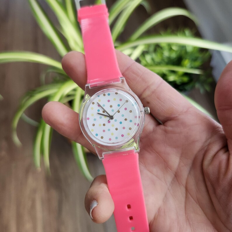 May include: A clear plastic watch with a white face and colorful polka dots. The watch has a bright pink silicone band.