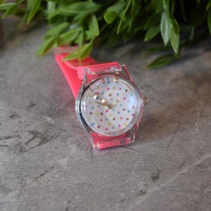 May include: A pink and clear plastic watch with a white face and colorful polka dots. The watch has a pink silicone band.
