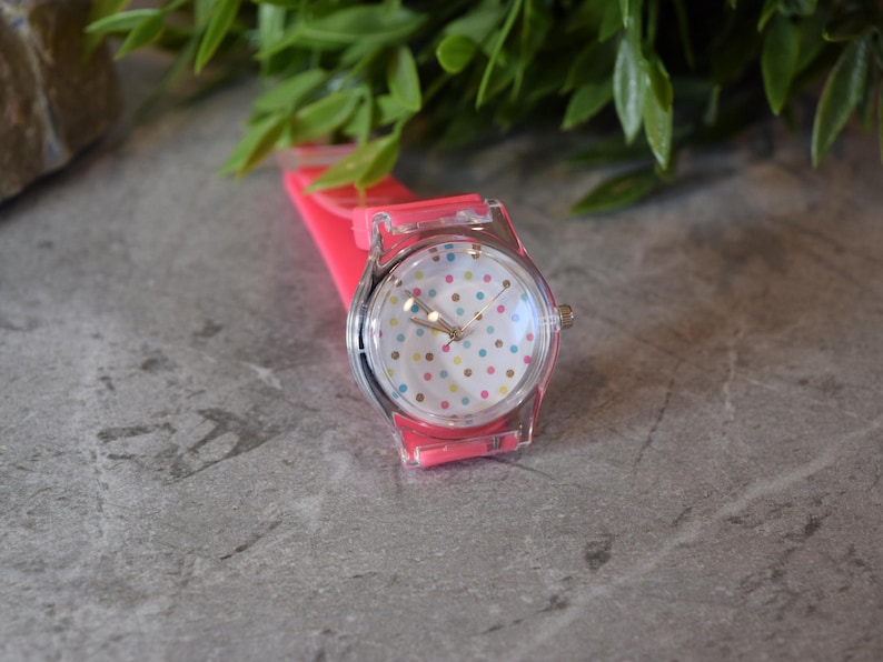May include: A pink and clear plastic watch with a white face and colorful polka dots. The watch has a pink silicone band.