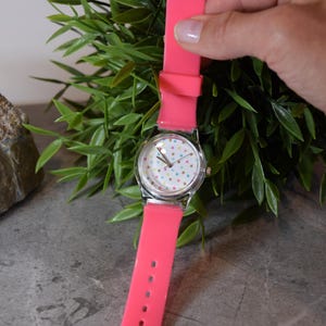 May include: A pink silicone wristwatch with a white face and colorful polka dots. The watch has a silver case and a silver band.