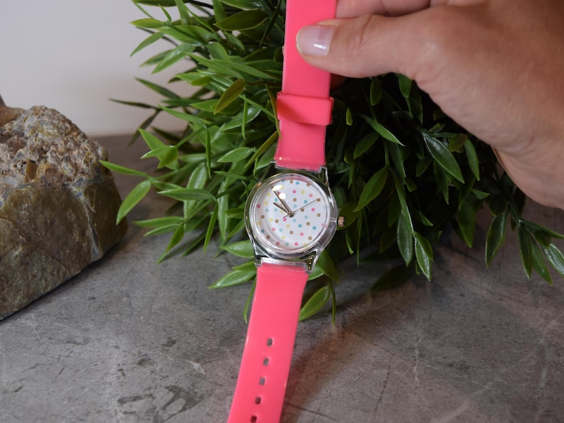 May include: A pink silicone wristwatch with a white face and colorful polka dots. The watch has a silver case and a silver band.