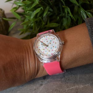 May include: A clear plastic watch with a pink silicone band. The watch face has a white background with colorful polka dots.