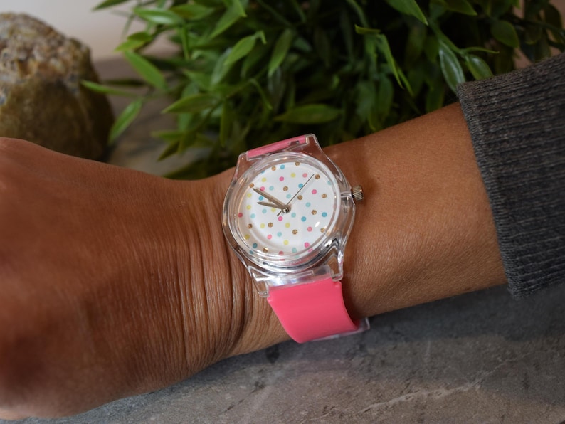 May include: A clear plastic watch with a pink silicone band. The watch face has a white background with colorful polka dots.