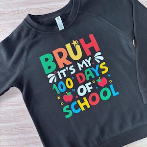 May include: Black sweatshirt featuring the phrase "BRUH IT'S MY 100 DAYS OF SCHOOL" in a vibrant, multi-coloured design. The graphic includes stars and apples. The sweatshirt has a crew neck.