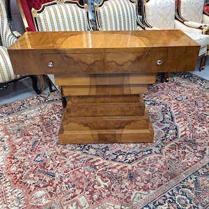 May include: A wooden console table with a stepped base and a single drawer. The table is made of light brown wood with a glossy finish. The drawer has a small, round, silver knob. The table is placed on a patterned rug. The table's dimensions are approximately 150 cm long.