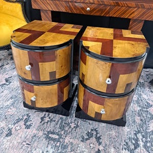 Op de afbeelding: Twee nachtkastjes in Art Deco-stijl met elk twee lades. Ze hebben een geometrisch ontwerp met lichte en donkere houten panelen, zwarte randen en zilveren ladeknopen. De nachtkastjes staan op een patroonkleed.