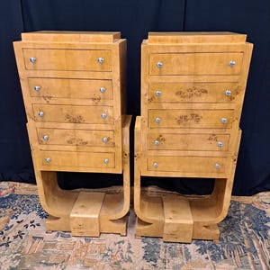 Op de afbeelding: Twee houten dressoirs in Art Deco-stijl, elk met zes lades en zilveren knoppen. De lichte houtnerf wordt geaccentueerd door een gebogen voet. De lades hebben decoratieve bloempatronen. De dressoirs zijn ongeveer 91 cm hoog.