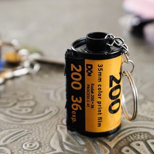 Op de afbeelding: Een zwarte en gele filmrol sleutelhanger met de tekst "200 36 exp." en "35mm color print film" erop gedrukt. De sleutelhanger is bevestigd aan een zilveren sleutelring.