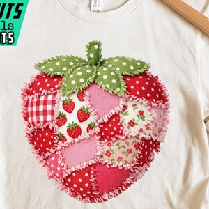 Puede incluir: Camiseta color crema con un diseño de fresa en patchwork. La fresa está compuesta por varios patrones de tela en rojo, rosa y verde, incluyendo lunares, cuadros vichy y estampados florales. La camiseta se muestra en una percha de madera.