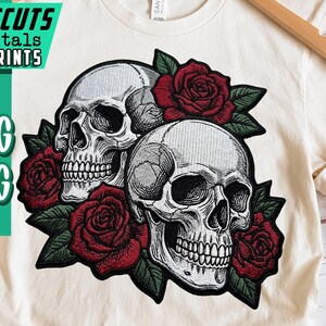 May include: Cream-colored t-shirt featuring a graphic of two skulls surrounded by red roses and green leaves. The shirt also has the text "ARTCUTS DIGITALS & PRINTS" and "PNG JPG" in a green and white design.