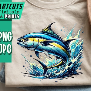 Puede incluir: Una camiseta beige con una vibrante ilustración de un pez azul y amarillo saltando del agua azul. La camiseta está doblada sobre unos vaqueros azules. Un sombrero marrón claro es visible en el fondo. El texto "ARTCUTS DIGITALS & PRINTS" está a la izquierda.