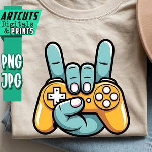 May include: A beige t-shirt with a graphic of a hand making a rock and roll gesture, holding a yellow video game controller. The hand is light blue with white accents. The text "ARTCUTS DIGITALS & PRINTS" is in the upper left corner.