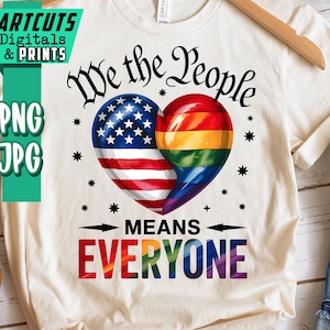 Puede incluir: Camiseta color crema con el texto "We the People Means Everyone". El diseño presenta un corazón dividido en dos mitades: una con la bandera estadounidense y la otra con los colores del arcoíris de la bandera del orgullo LGBTQ+. La camiseta está colgada en una percha de madera.