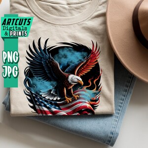 May include: A beige t-shirt featuring a detailed graphic of a bald eagle with outstretched wings, perched atop an American flag. The eagle has brown, red, and white feathers. The shirt is folded on top of blue jeans, with a tan hat visible.