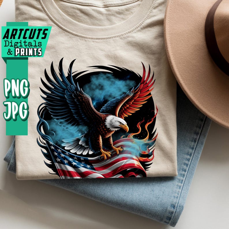 May include: A beige t-shirt featuring a detailed graphic of a bald eagle with outstretched wings, perched atop an American flag. The eagle has brown, red, and white feathers. The shirt is folded on top of blue jeans, with a tan hat visible.
