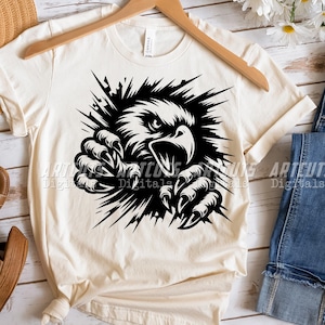 Pode incluir: Uma camiseta branca com um gr&aacute;fico preto de uma &aacute;guia rompendo uma parede. A cabe&ccedil;a e as garras da &aacute;guia s&atilde;o vis&iacute;veis.