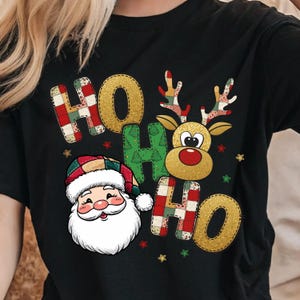 May include: Black t-shirt with a Christmas-themed design. The design features the words "HO HO" in a patchwork style, a Santa Claus face, and a reindeer with a red nose. Small star accents are also included in the festive design.