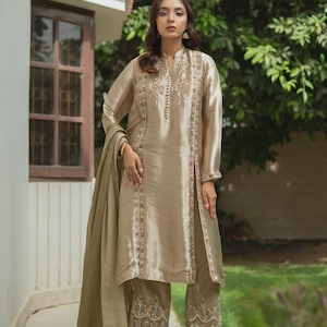 May include: A beige silk suit with intricate embroidery. The long-sleeved tunic and matching pants feature detailed embellishments along the seams and hems. A matching scarf drapes over the shoulder. The outfit is worn with high-heeled sandals.