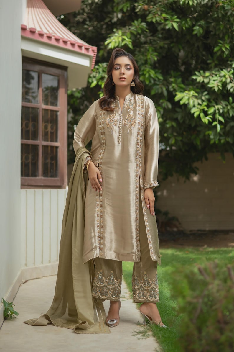 May include: A beige silk suit with intricate embroidery. The long-sleeved tunic and matching pants feature detailed embellishments along the seams and hems. A matching scarf drapes over the shoulder. The outfit is worn with high-heeled sandals.