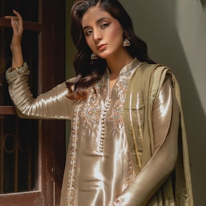 May include: A shimmering, champagne-colored silk ensemble featuring intricate embroidery and beadwork. The outfit includes a long-sleeved tunic with a mandarin collar and a matching scarf. The embroidery incorporates gold, teal, and cream-colored threads. The fabric has a subtle sheen.