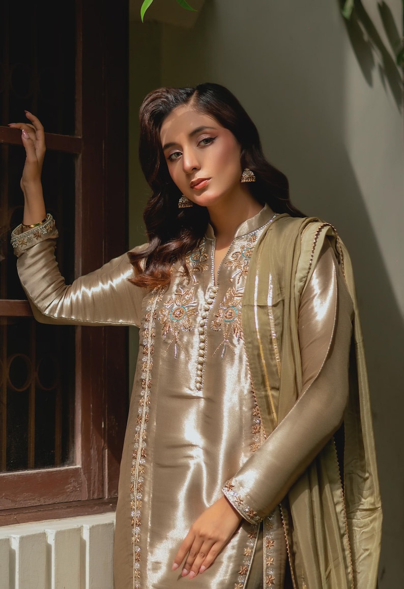 May include: A shimmering, champagne-colored silk ensemble featuring intricate embroidery and beadwork. The outfit includes a long-sleeved tunic with a mandarin collar and a matching scarf. The embroidery incorporates gold, teal, and cream-colored threads. The fabric has a subtle sheen.