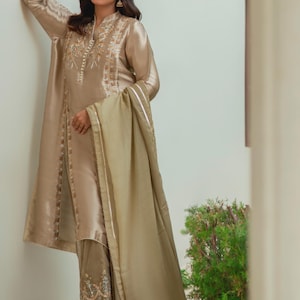 May include: A beige silk ensemble featuring a long tunic with intricate embroidery around the neckline and down the front. The outfit includes matching pants with detailed embroidery at the ankles and a flowing scarf. The outfit is a formal wear.