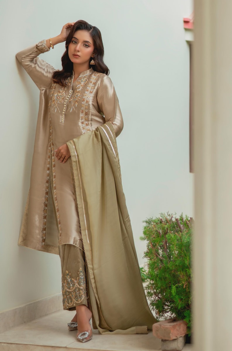 May include: A beige silk ensemble featuring a long tunic with intricate embroidery around the neckline and down the front. The outfit includes matching pants with detailed embroidery at the ankles and a flowing scarf. The outfit is a formal wear.