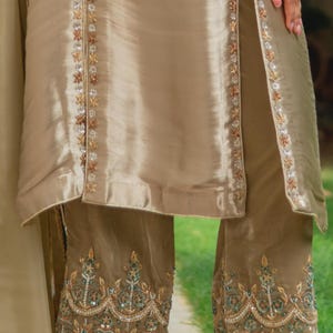 May include: A two-piece beige outfit with intricate embroidery. The top has a split front with floral embellishments. The pants feature detailed embroidery along the hemline, with gold, green, and silver accents. The fabric has a subtle sheen.